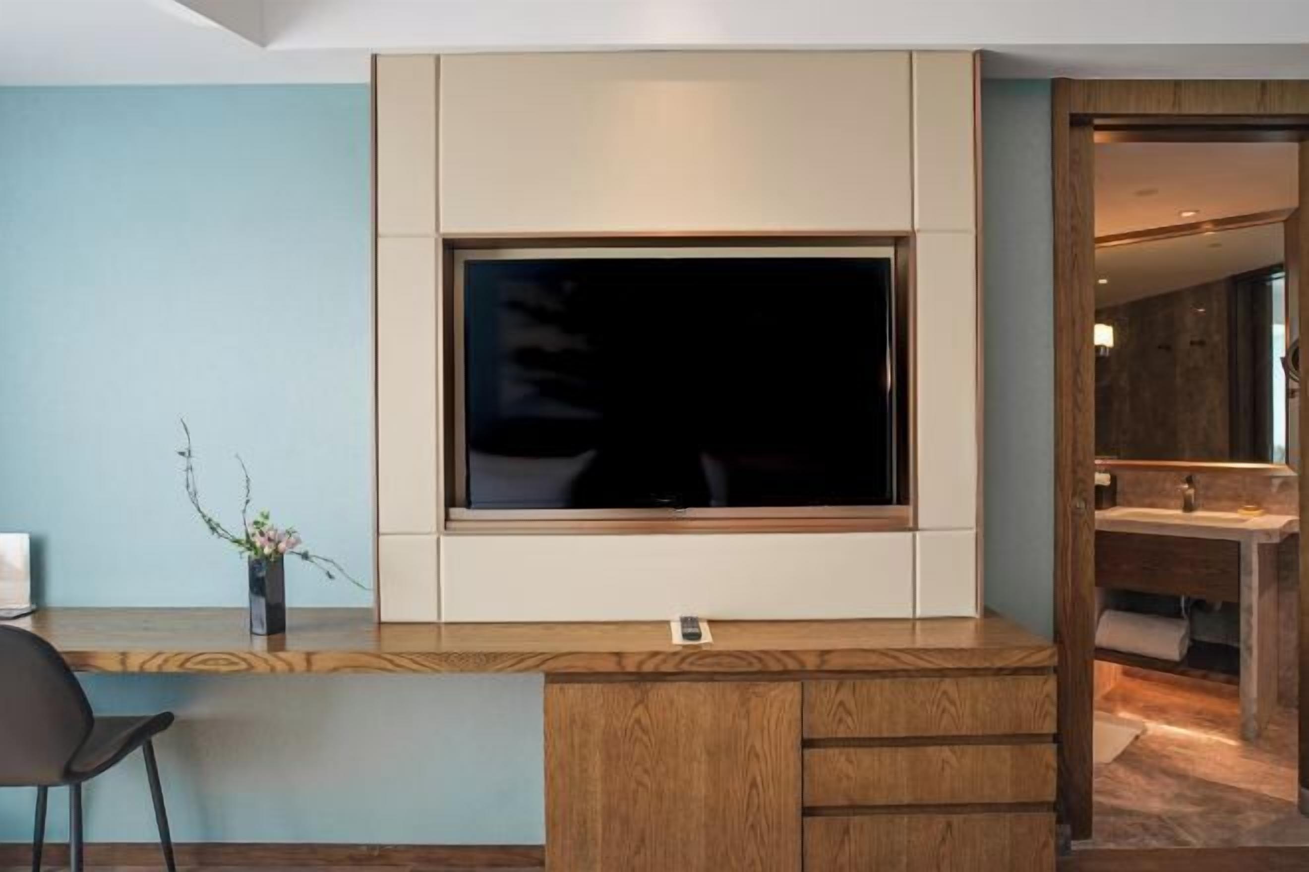 Una habitación moderna con un televisor grande montado en la pared, un escritorio de madera con una silla, y un baño visible a través de una puerta abierta.