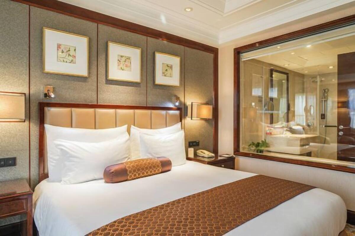 Une chambre d’hôtel avec un grand lit, une tête de lit en bois, des œuvres d’art encadrées accrochées au mur et une grande fenêtre donnant sur la salle de bain.