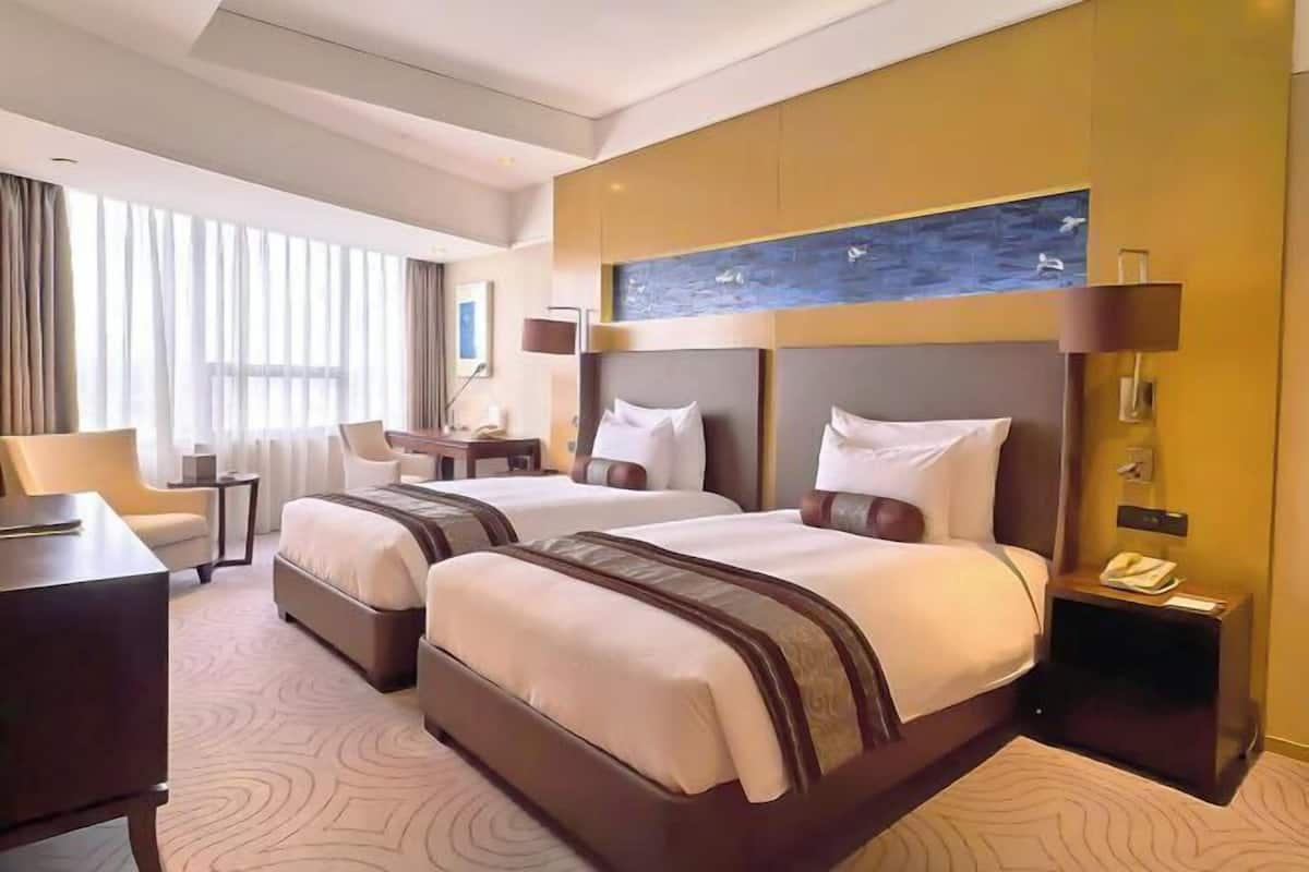 Une chambre d’hôtel avec deux lits, un bureau, une chaise et une grande fenêtre avec des rideaux.