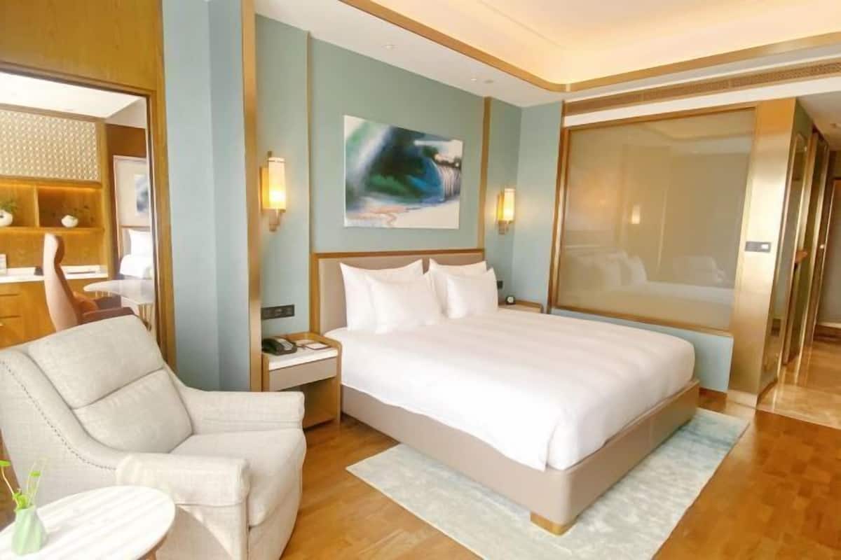 Une chambre d’hôtel avec un grand lit, un coin salon comprenant une chaise et une petite table, une table de chevet avec une lampe, et un tableau accroché au mur.