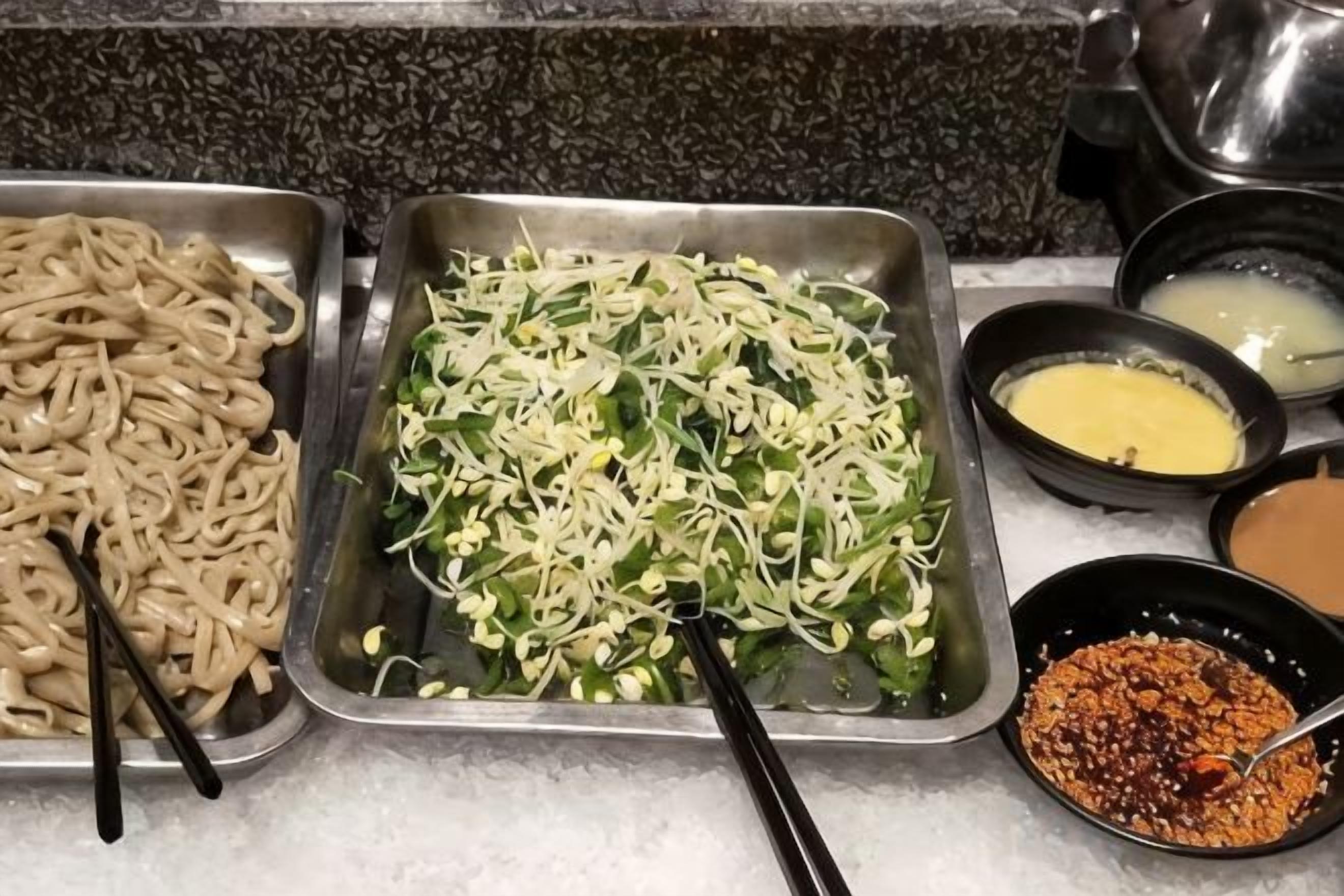 Un bufé con diferentes opciones de comida, incluyendo fideos, vegetales picados y salsas.