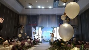 Ein Festsaal mit gedeckten Tischen für eine festliche Veranstaltung, eine Bühne mit Luftballons und ein großer Eingangsbereich.