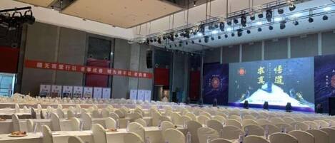 Un salón de conferencias con filas de sillas, un escenario con una pantalla grande y pancartas en las paredes.