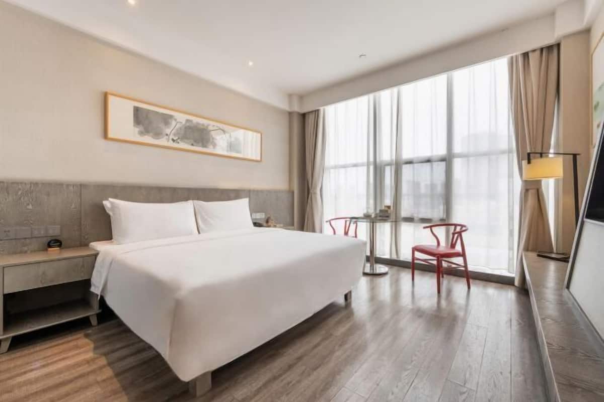 A hotel room with a bed, a nightstand, a chair, a table, and a large window with curtains.