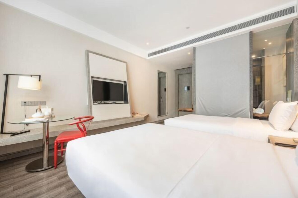 A hotel room with a large bed, a desk with a chair, a TV, and a glass partition.