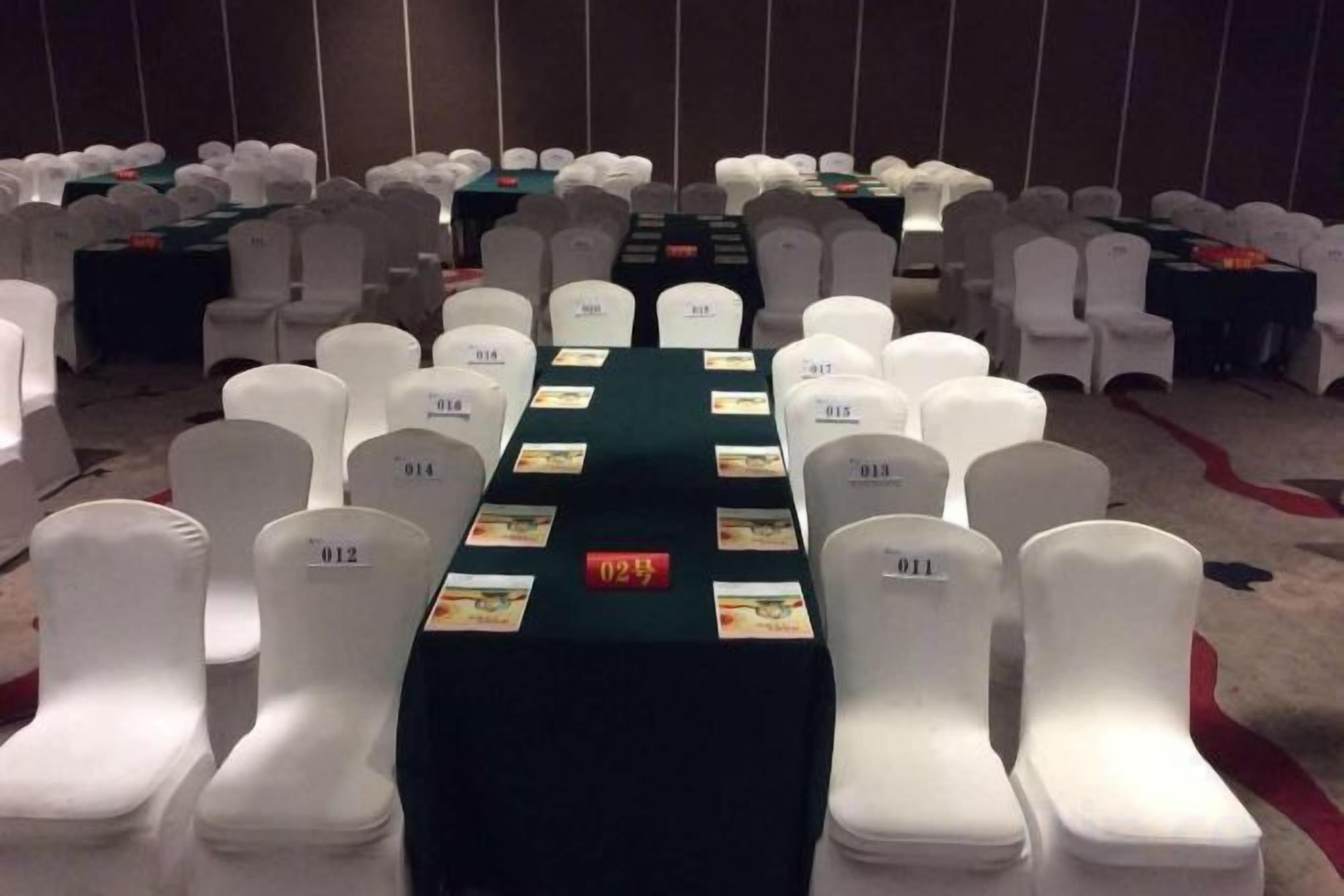 A conference room with rows of white chairs, a long table with name cards, and a carpeted floor.