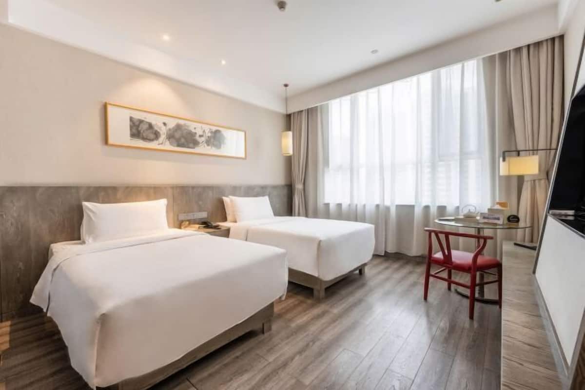 A hotel room with two beds, a desk, a chair, a window with curtains, and a framed picture on the wall.
