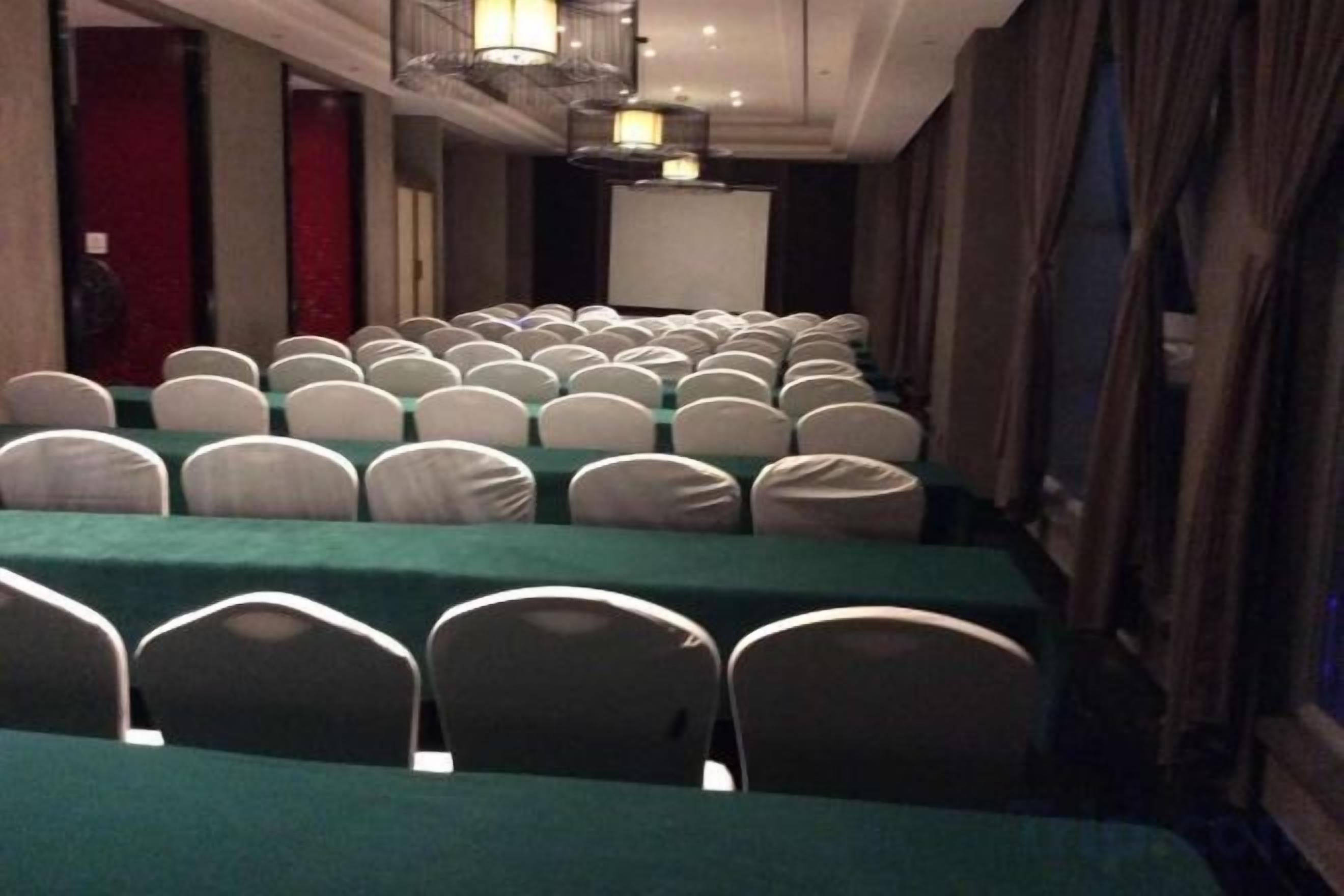 Un salón de conferencias con filas de mesas y sillas, una pantalla de proyector y un escenario.