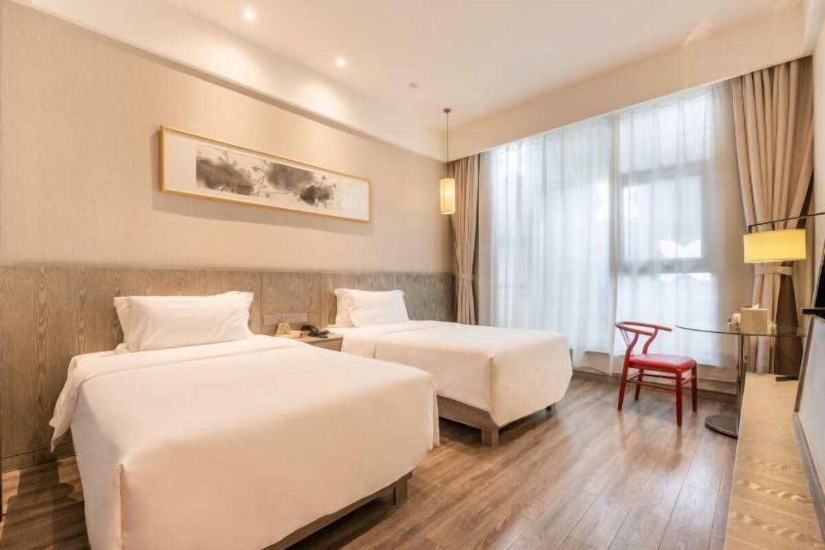 A hotel room with two beds, a desk, a chair, a lamp, and a large window with curtains.