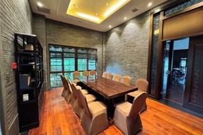 A conference room with a long table and chairs, a bookshelf, and a view through a window.