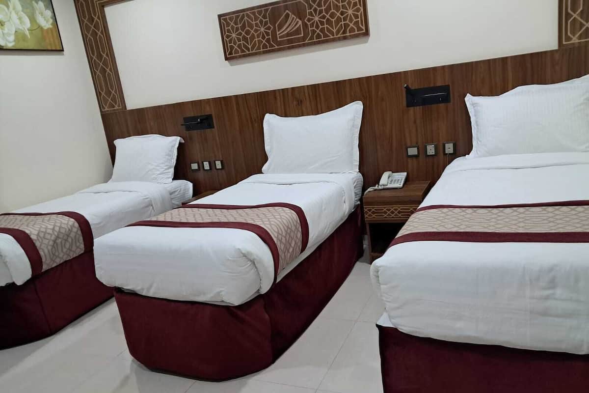 A hotel room with three single beds, each with a red bed skirt and white bedding. There is a wooden headboard with a geometric design, a bedside table with a phone, and a wall-mounted light switch.