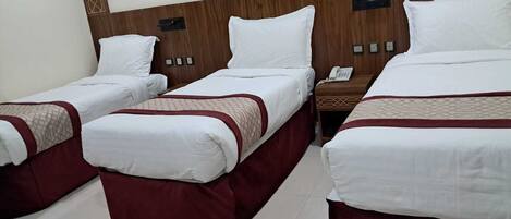 Habitación de hotel con tres camas individuales, cada una con falda roja y ropa de cama blanca. Tiene un cabecero de madera con diseño geométrico, una mesita de noche con teléfono y un interruptor de luz en la pared.