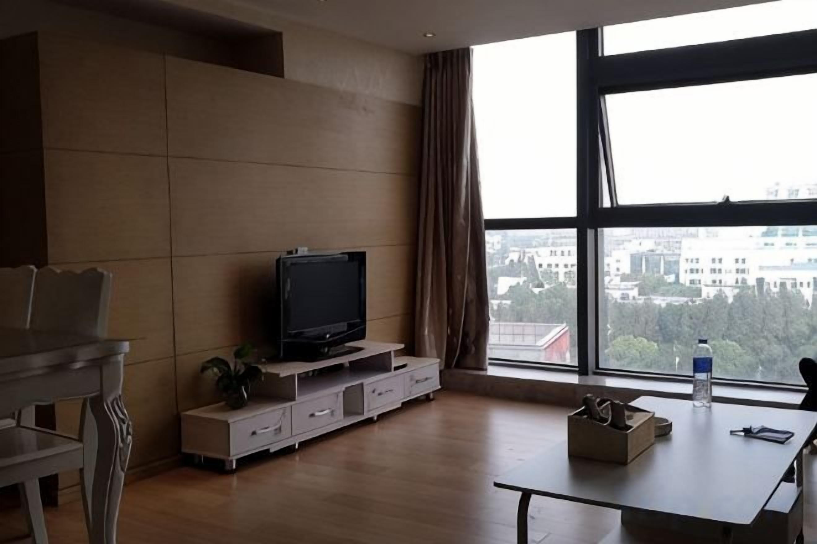 Uma sala de estar moderna com televisão de tela plana sobre um suporte de madeira, uma mesa de jantar com cadeiras e uma grande janela com vista para a cidade.