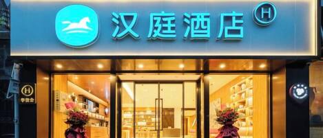 A hotel entrance with a neon sign, glass doors, and two flower arrangements on either side.