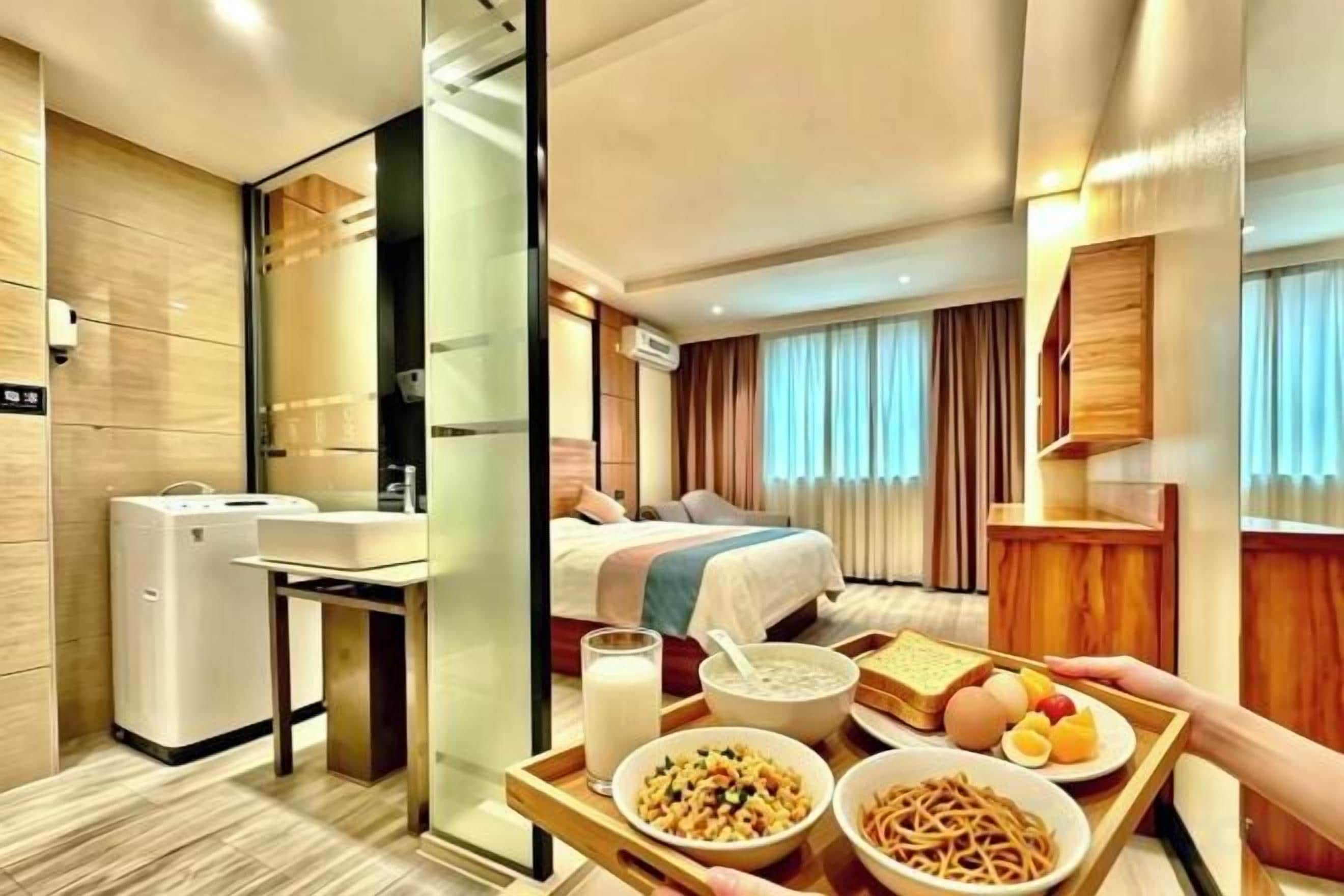 Quarto de hotel com cama, mesa de cabeceira, divisória de vidro, mini-geladeira e bandeja com itens de café da manhã.