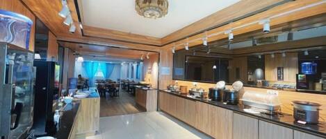 A modern hotel lobby with a long reception desk, a dining area in the background, and a large chandelier.
