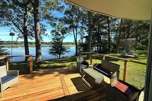 Una terraza de madera con mobiliario de mimbre que da a un lago, rodeada de árboles.