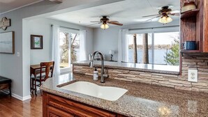 A kitchen with granite countertops, a built-in sink, wooden cabinets, a stone backsplash, and a large window overlooking a body of water.