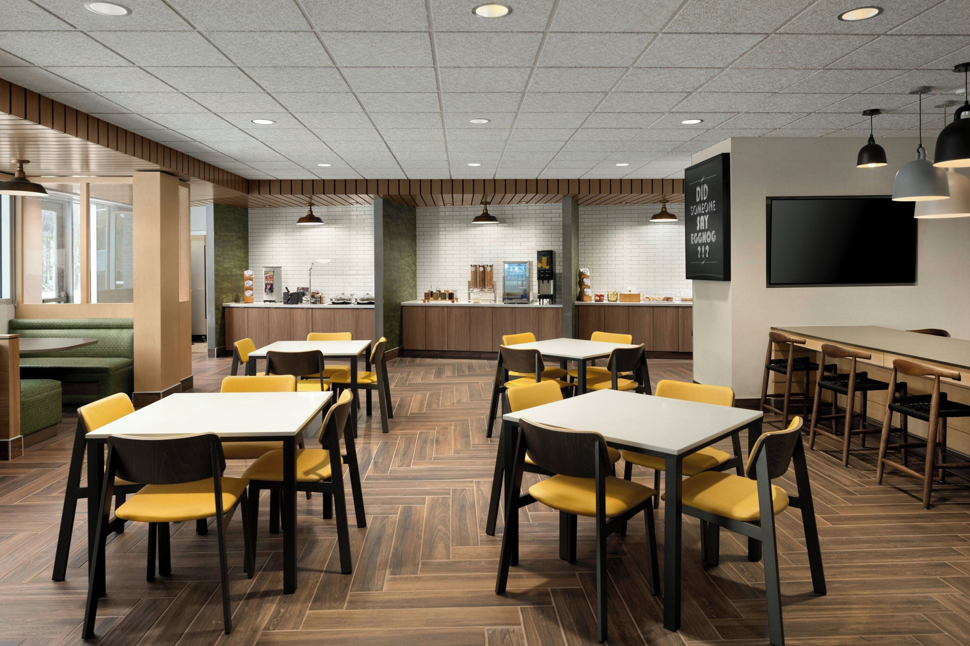 Un espace repas moderne avec un plancher en bois, des chaises jaunes et des tables blanches. On y trouve également un bar et un grand téléviseur à écran plat fixé au mur.
