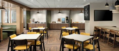 Un espace repas moderne avec un plancher en bois, des chaises jaunes et des tables blanches. On y trouve également un bar et un grand téléviseur à écran plat fixé au mur.