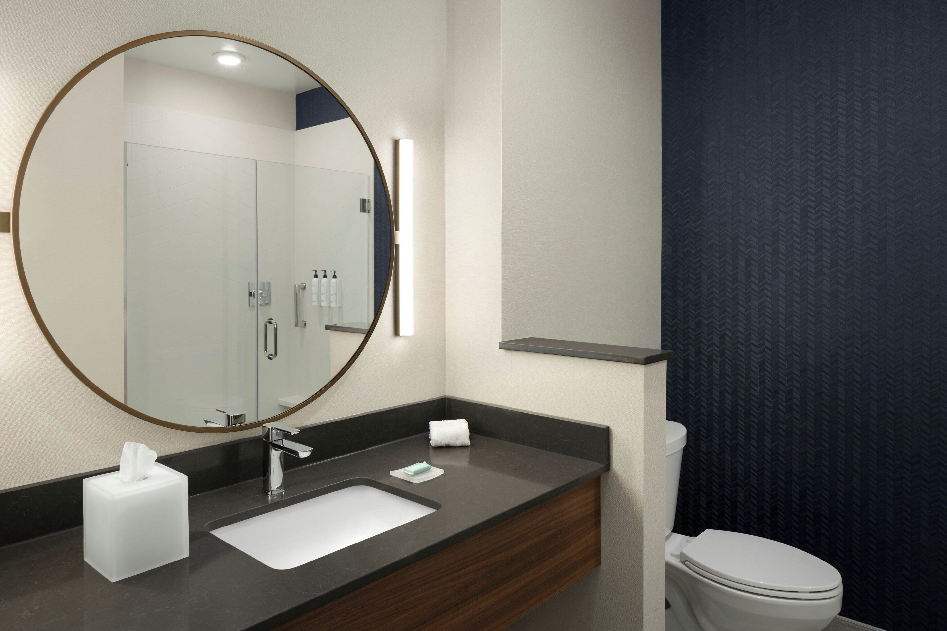 A modern bathroom with a round mirror, a sink, a toilet, and a towel dispenser.