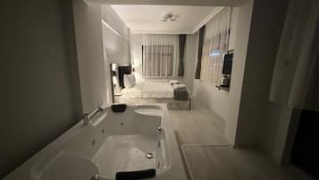A modern hotel room with a large bathtub, a bed with a white comforter, and a television on a shelf.