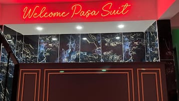 Un mostrador de recepción con un letrero de neón que dice "Welcome Paso Suit" y una gran pared oscura, similar a mármol, en el fondo.
