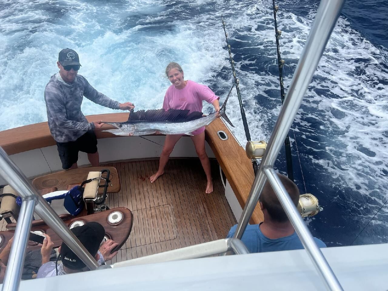 A boat deck with a wooden floor, a table with chairs, and a person holding a fishing rod.