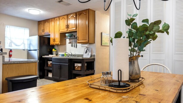 Una cocina con armarios de madera, una mesa de comedor de madera, un florero con hojas verdes, saleros y pimenteros, y un dispensador de toallas de papel.