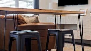 A modern living room with a wooden coffee table, two black stools, a brown sofa, and a flat-screen TV mounted on the wall.