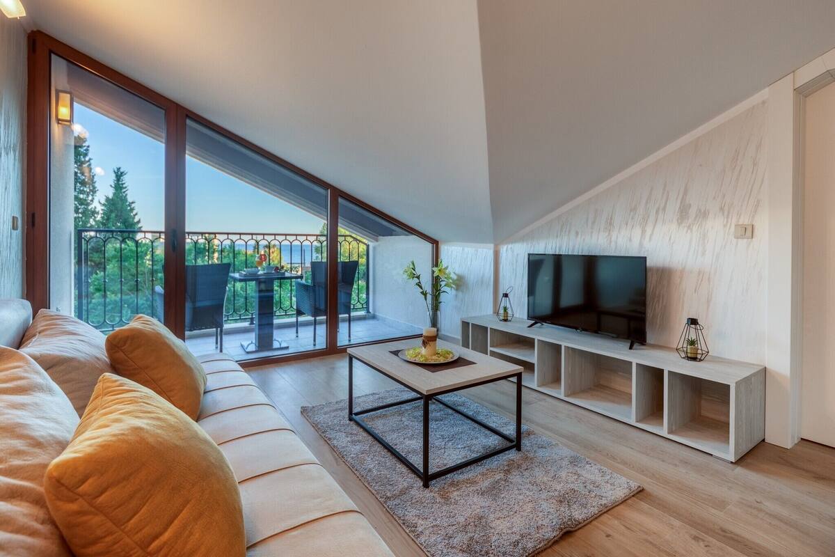 A modern living room with a large sofa, a coffee table, and a TV on a wooden stand. There is a balcony with a view of trees and a railing.