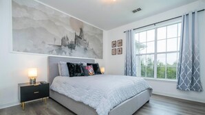 Ein Schlafzimmer mit einem großen Bett, einem Nachttisch mit Lampe, einem Fenster mit Vorhängen und einer Wand mit einem großen Gemälde.