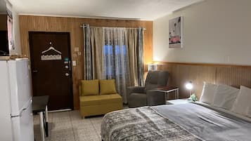 Habitación de hotel con cama, sofá, sillón, mesita, lámpara y puerta con perchero.