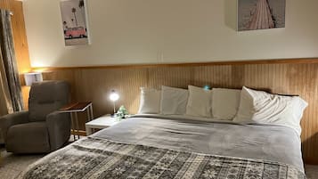 A hotel room with a wooden headboard, a bed with patterned bedding, a chair, a nightstand with a lamp, and two wall art pieces depicting a car and a beach scene.