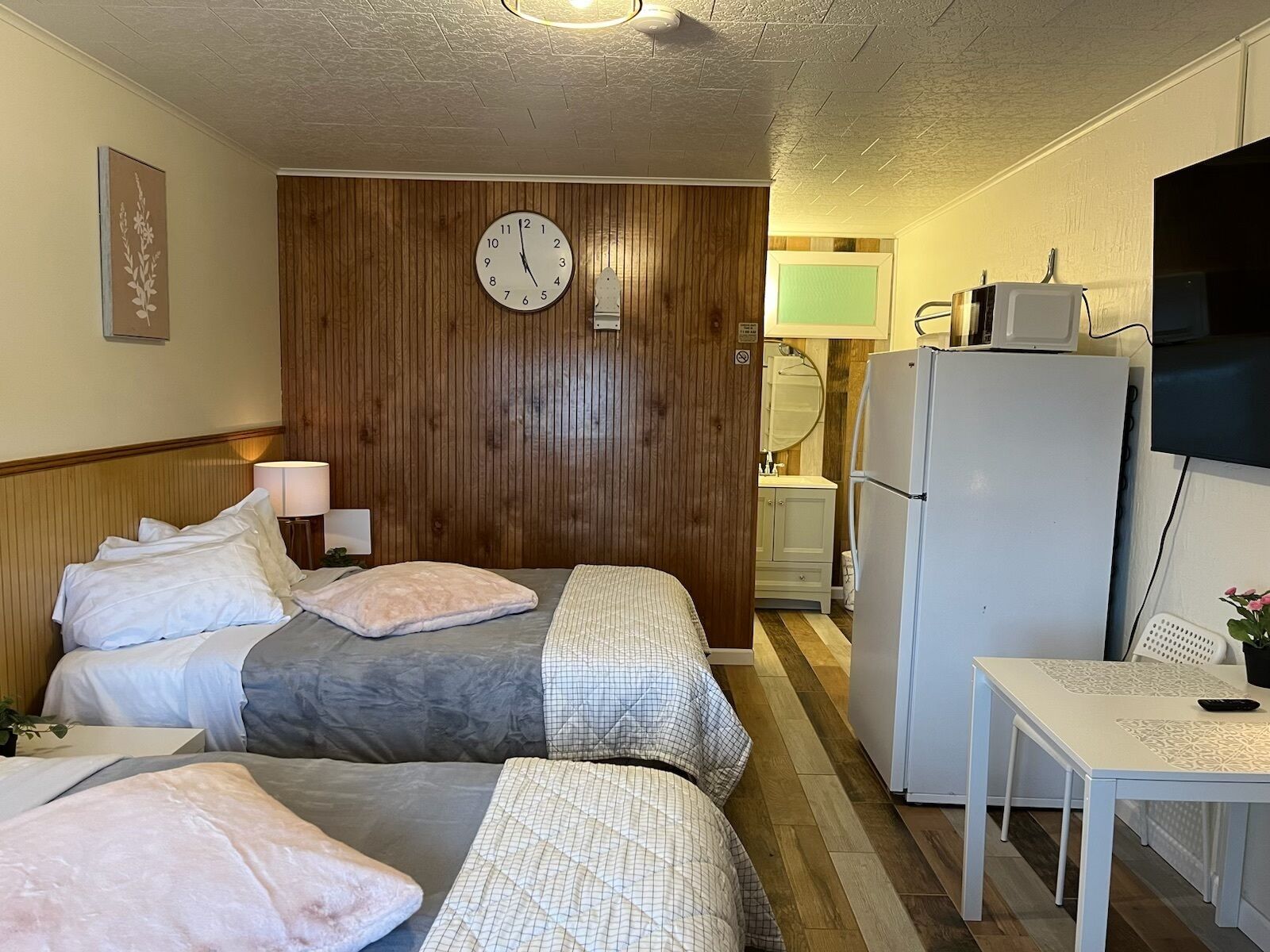 Une chambre d’hôtel avec deux lits, un mur recouvert de panneaux de bois, une horloge, un réfrigérateur, une télévision et une petite table avec une chaise.