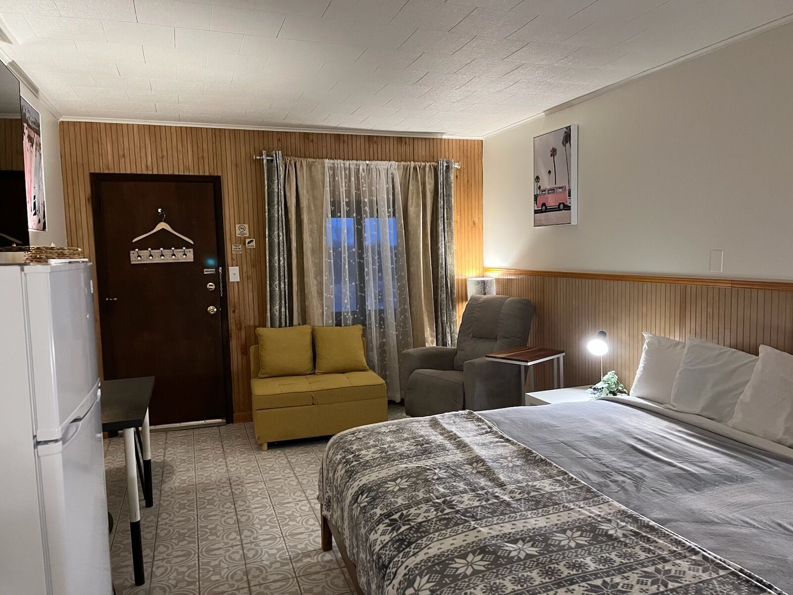 A hotel room with a bed, a sofa, a chair, a desk, a lamp, and a door with a coat rack.