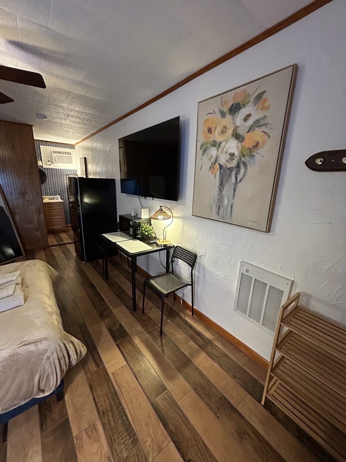 Quarto compacto com uma cama, escrivaninha, cadeira, televisão e um quadro.