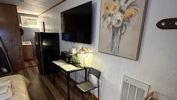 Quarto compacto com uma cama, escrivaninha, cadeira, televisão e um quadro.