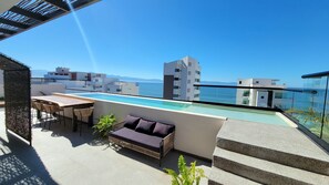 Eine Dachterrasse mit Pool, Sitzgelegenheiten im Freien und Blick auf das Meer und die Gebäude.