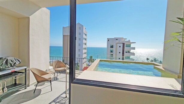 A balcony with a view of the ocean, a pool, and a dining area with chairs and a table.