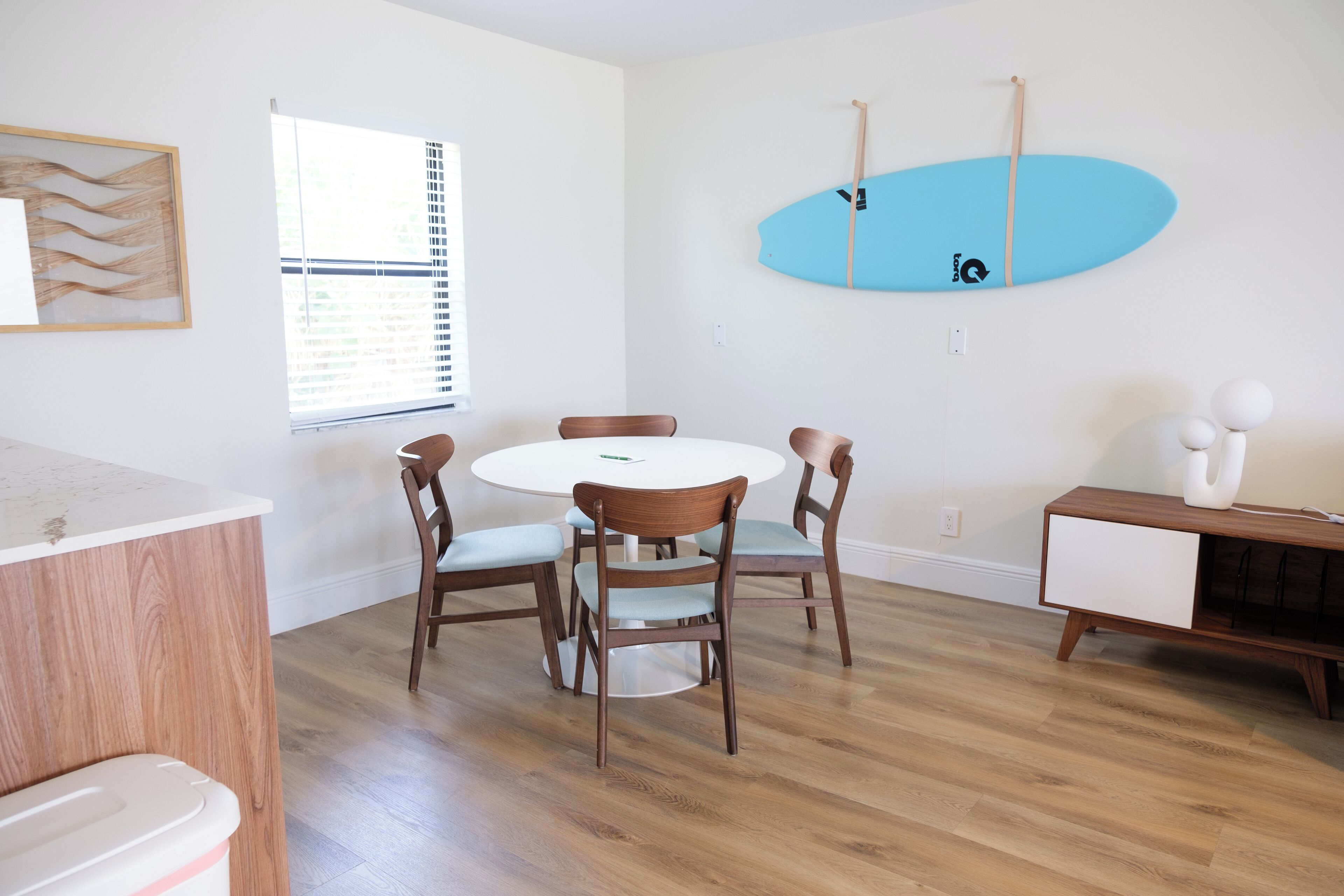 Un comedor con una mesa redonda y sillas, una tabla de surf en la pared y un armario con tapa blanca.