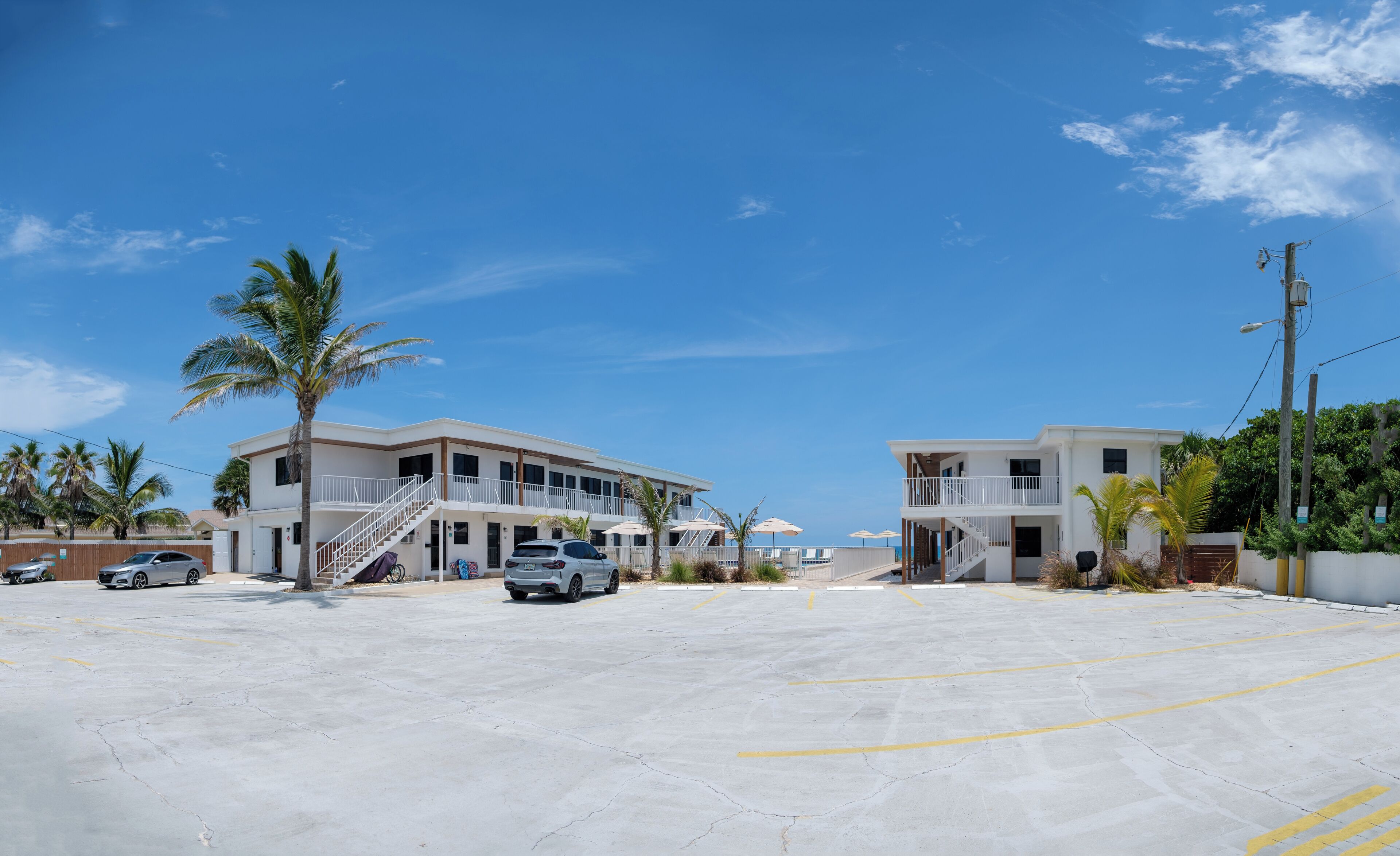 A beachfront hotel with two-story buildings, a parking area, and palm trees.