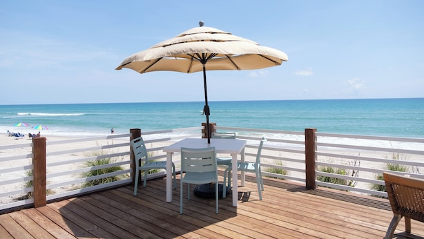 A wooden deck with a table and chairs, an umbrella, and a view of the beach and ocean. - Melbourne Beach Resort & Surf Club  (Melbourne Beach)