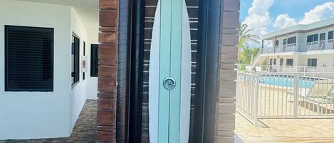 A building entrance with a surfboard-shaped door, brick pillars, and a pool area visible in the background.