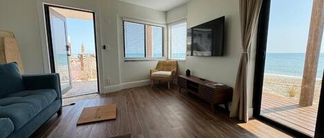 A modern living room with a wooden floor, a blue sofa, a wooden coffee table, and a view of the beach through large windows.