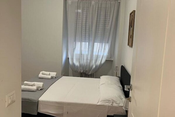 A small, neatly arranged bedroom with a single bed, a bedside table, and a window with sheer curtains.