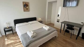Una camera d'albergo con un letto grande, una scrivania, una sedia, una TV e un tavolino con una lampada.