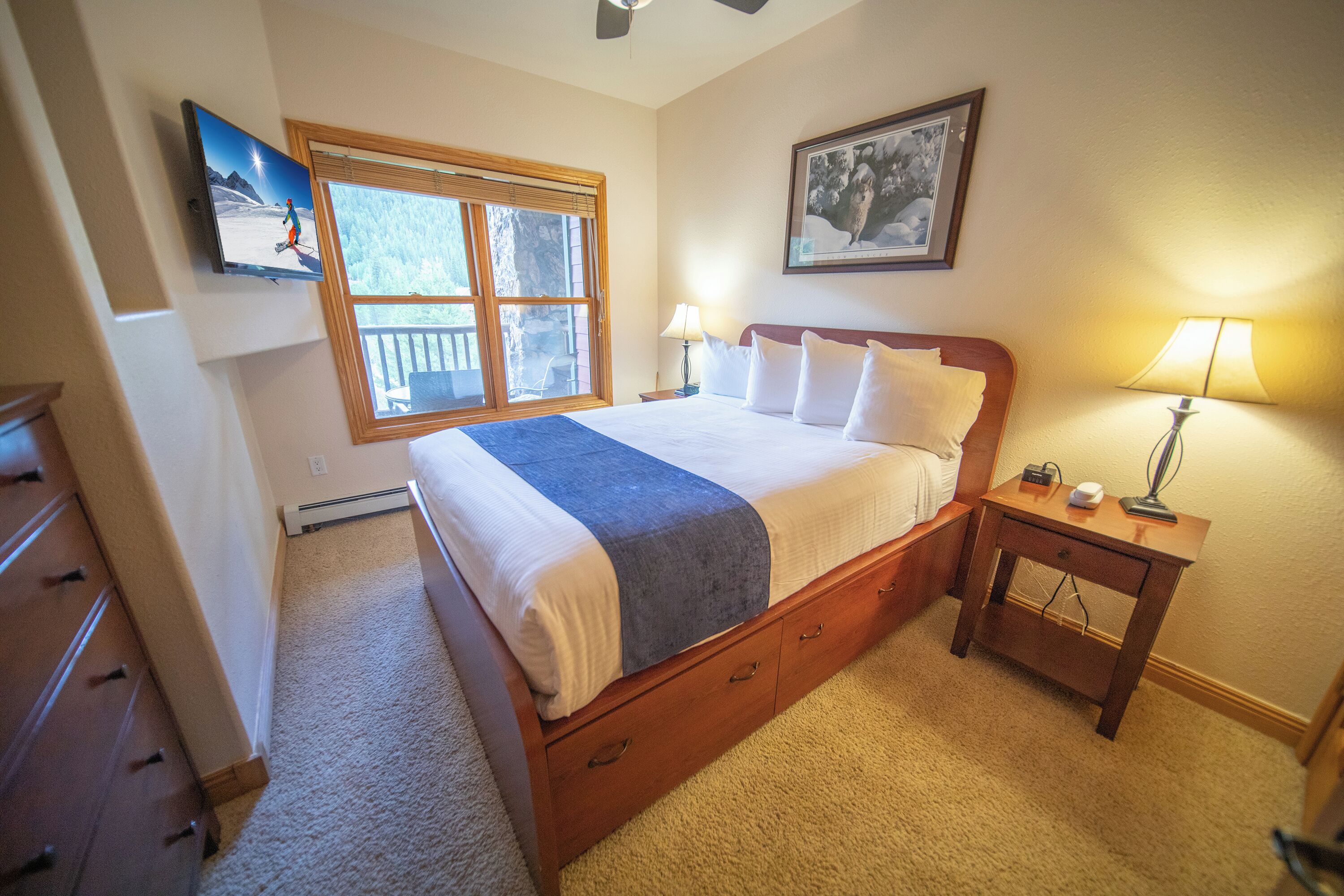 A bedroom with a large bed, a nightstand, a dresser, a window with a view, and a TV mounted on the wall.