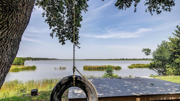 Une balançoire faite d’un pneu suspendu à un arbre, une terrasse en bois et un lac paisible, avec des roseaux au premier plan.