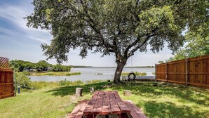 Une table de pique-nique se trouve dans une zone herbeuse, avec un lac et une clôture en bois en arrière-plan.